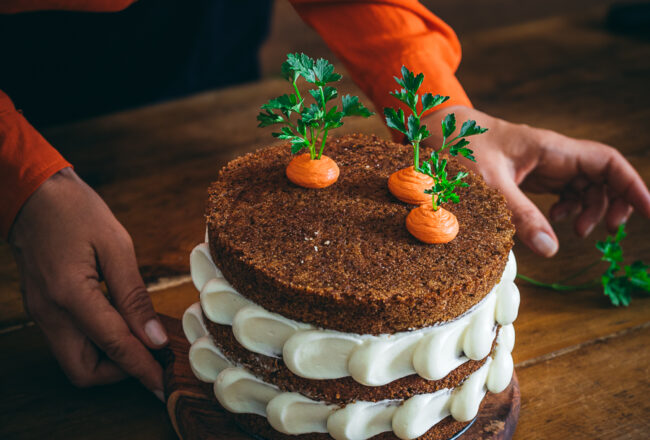 torta di carote e mele a strati con decorazione di frosting bianco e finte carote su tavolo legno